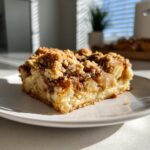 Ein Stück saftiger Apfel-Zimt-Streuselkuchen auf einem weißen Teller, mit knusprigen Streuseln und Apfelstücken.