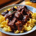 Ein Teller mit Gulasch mit Nudeln oder Spätzle, bedeckt mit einer reichhaltigen, dunklen Soße und zarten Fleischstücken.