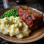 Ein saftiges Stück Hackbraten mit Kartoffelpüree und Erbsen auf einem Teller, ein echtes Wohlfühlgericht.