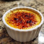 Nahaufnahme einer Crème Brûlée mit goldbrauner, knackiger Zuckerkruste im weißen Förmchen.