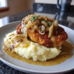 Ein goldbraunes Senf-Zwiebel-Schnitzel serviert auf cremigem Kartoffelbrei, übergossen mit einer würzigen Sauce und garniert mit karamellisierten Zwiebeln.