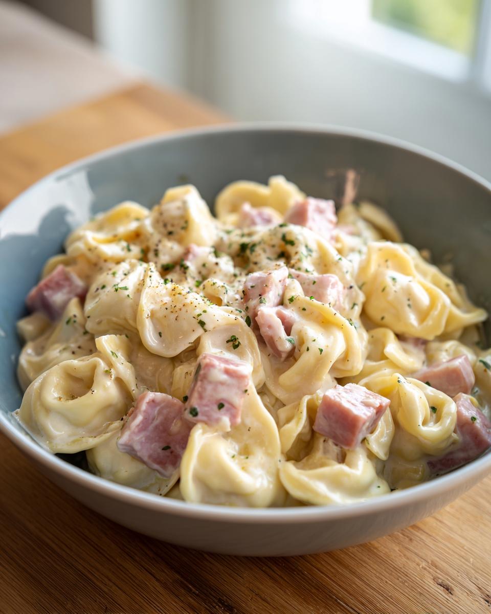 Eine Schüssel voller cremiger Tortellini in Schinken-Sahne-Sauce, garniert mit Petersilie und Pfeffer.