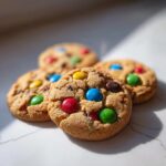 Vier frisch gebackene, weiche Smarties Cookies mit bunten Schokolinsen liegen auf einer hellen Oberfläche.