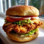 Nahaufnahme eines saftigen Crispy Chicken Burger mit knusprigem Hähnchen, Salat, Tomate und Zwiebelringen.