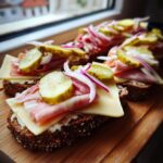 Mehrere Scheiben dunkles Brot belegt mit Frischkäse, Käse, Schinken, roten Zwiebelringen und sauren Gurken für eine schnelle Brettljause.