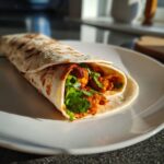 Nahaufnahme eines fertig gerollten Chicken-Tikka-Wrap, gefüllt mit würzigem Hähnchen und frischem Koriander, auf einem weißen Teller.