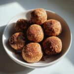 Nahaufnahme von mehreren Apfel-Zimt-Protein-Bällchen, bestäubt mit Zimt, in einer hellen Schale.