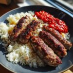 Nahaufnahme von saftigen, gegrillten Ćevapčići mit weißem Reis und einem Klecks rotem Ajvar in einer dunklen Schüssel.