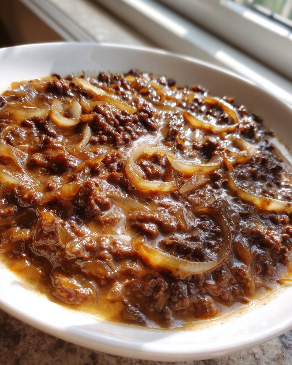 Nahaufnahme von saftigen Frikadellen in einer dunklen, reichhaltigen Zwiebelsauce mit sichtbaren Zwiebelringen.