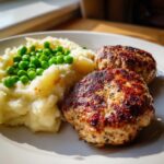 Zwei goldbraun gebratene Frikadellen neben cremigem Kartoffelpüree und leuchtend grünen Erbsen.