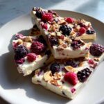 Mehrere Stücke Frozen-Joghurt-Rinde mit Beeren und Mandeln auf einem weißen Teller.