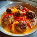 Nahaufnahme von saftigen Hackbällchen in Paprikarahm mit roten und gelben Paprikastreifen in einer Schüssel.