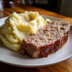 Ein Anschnitt von saftigem Hackbraten neben einer großen Portion cremigem Kartoffelpüree auf einem Teller.