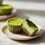 Zwei cremige Matcha-Cheesecake-Bites, einer davon angeschnitten, um den Boden zu zeigen.