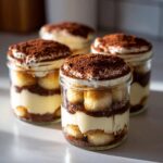 Vier fertige Mini-Tiramisu-Törtchen im Glas, geschichtet mit Löffelbiskuits, Creme und reichlich Kakaopulver.