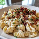 Nahaufnahme einer cremigen Nudelpfanne mit Frischkäse, garniert mit knusprigem Speck und Schnittlauch.