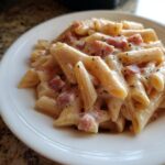 Nahaufnahme von Penne mit Schinken-Sahnesauce, bedeckt mit cremigem Käse und schwarzem Pfeffer.