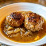 Nahaufnahme von zwei goldbraun gebratenen Frikadellen in einer reichhaltigen Zwiebelsauce.