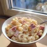 Nahaufnahme einer dampfenden Schüssel Schinken-Käse-Nudeln aus einem Topf mit cremiger Soße und Schinkenwürfeln.