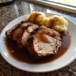 Teller mit aufgeschnittenem Schweinebraten, dunkler Soße und zwei Portionen Kartoffelklößen.