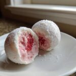 Zwei Strawberry-Mochi-Eis Kugeln auf einem Teller, eine davon aufgeschnitten, zeigt die cremige rosa Füllung und das rote Erdbeerherz.