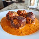 Nahaufnahme von vier saftigen Köfte, die in einer cremigen, orange-roten Tomaten-Sahnesauce liegen.