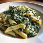Nahaufnahme von Penne Nudeln mit cremigem Spinat und Schmand in einem weißen Teller.