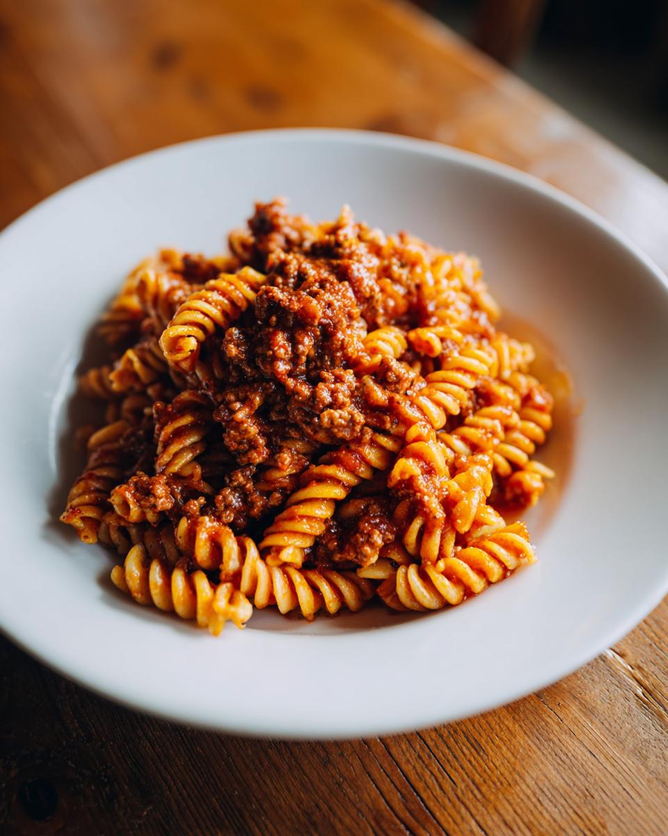 Nahaufnahme einer Portion Nudelpfanne mit Hackfleisch und Tomatenmark, serviert in einer weißen Schüssel auf Holz.